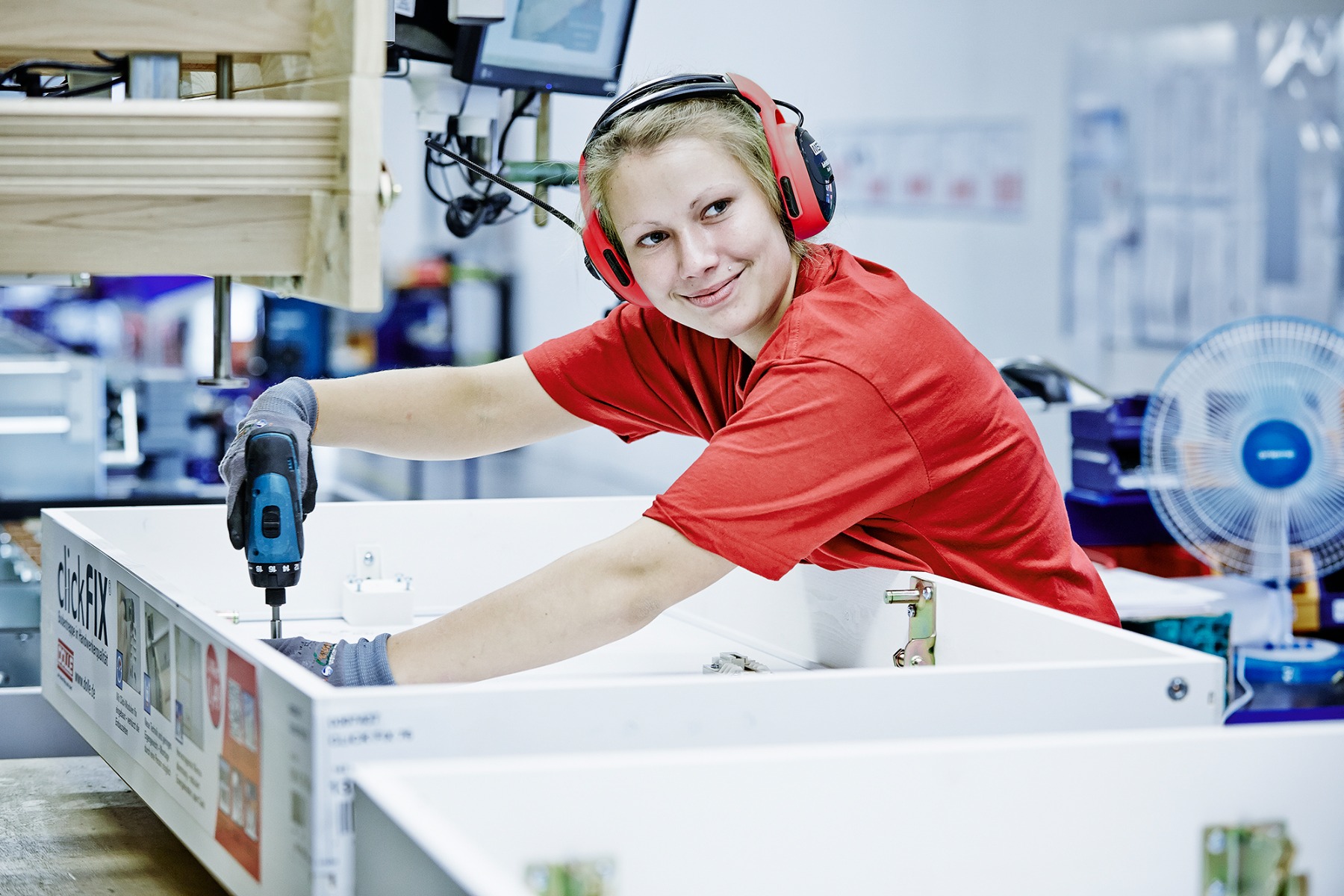 Fabrieksarbeider in een rood shirt die een dakkap monteert met een elektrische boormachine, terwijl hij handschoenen en gehoorbescherming draagt.