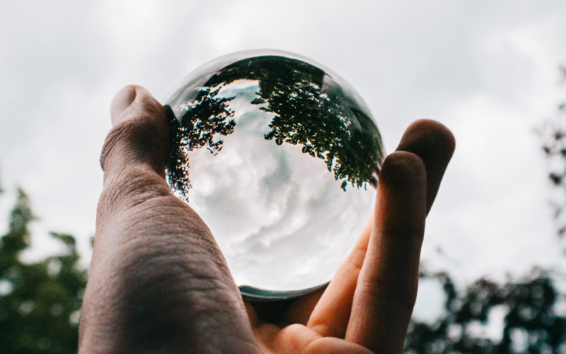 Hand die een reflecterende glazen bol vasthoudt, met een omgekeerd uitzicht op bomen en een bewolkte lucht.